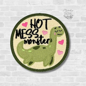 May include: Embroidered patch with the text "HOT MESS monster" and a green dinosaur illustration. The patch is round with a cream-colored background, pink hearts, and a dark green border. The dinosaur has a friendly expression.