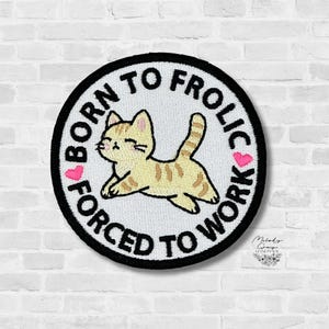May include: Embroidered patch with a black border and the text "BORN TO FROLIC, FORCED TO WORK" around a cartoon cat. The cat is tan with brown stripes and pink hearts are on either side of the cat.