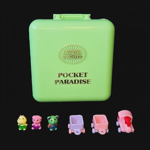 May include: A light green Pocket Paradise toy set. The set includes a rectangular case with the words "Small World" and "Pocket Paradise" in gold lettering. Also included are miniature toy figures and vehicles.