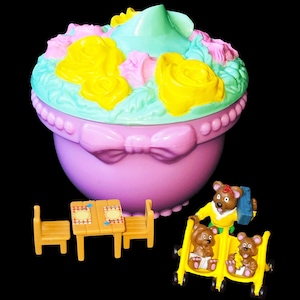 Puede incluir: Un juguete vintage con forma de cupcake con una parte superior floral en amarillo, rosa y verde. Incluye muebles en miniatura y figuras de osos, con un cochecito amarillo. El juguete está sobre un fondo negro.