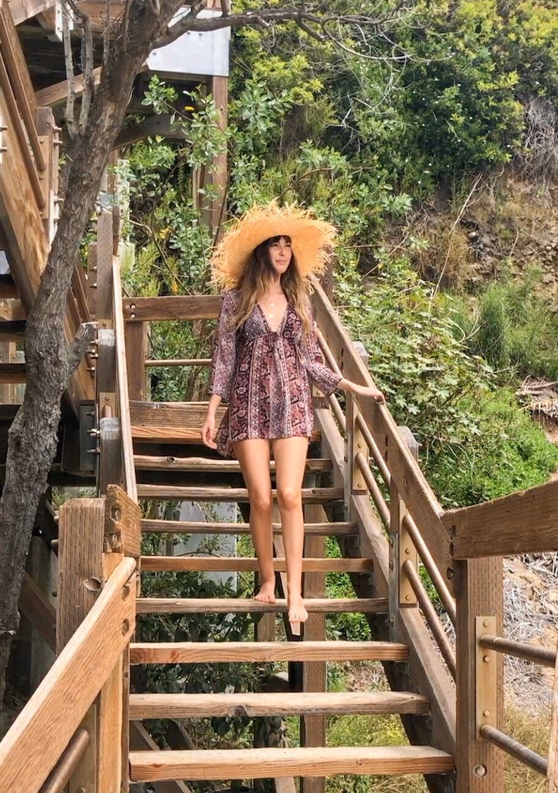 May include: A woman wearing a straw hat and a patterned dress walks down a wooden staircase. The staircase is surrounded by greenery.