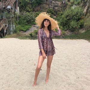 May include: A woman wearing a floral print dress and a wide-brimmed straw hat stands on a sandy beach.