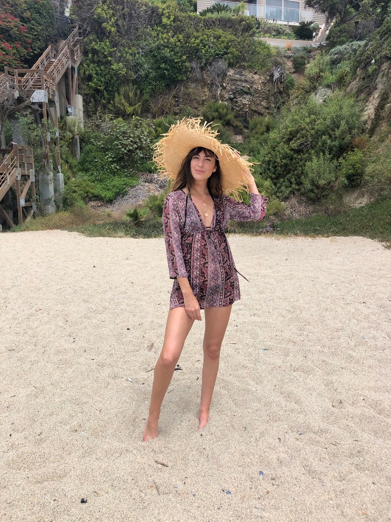 May include: A woman wearing a floral print dress and a wide-brimmed straw hat stands on a sandy beach.
