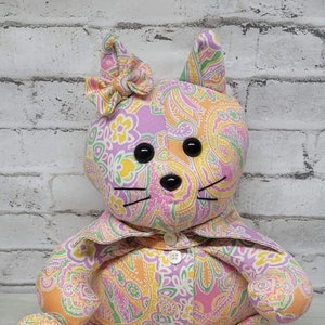 May include: A handmade cat-shaped stuffed animal made from patterned fabric in shades of pink, orange, yellow, and green. Features black button eyes, whiskers, and a bow. The word "grandma" and dates are embroidered on the feet.