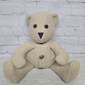 May include: A beige teddy bear with black button eyes and a black nose. The bear is wearing a beige sweater with two gold buttons. The bear is sitting with its legs spread out.