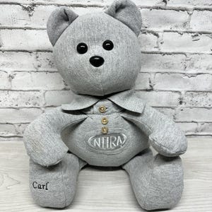 May include: A gray plush teddy bear wearing a gray collared shirt with a gold button and an embroidered NHRA logo. The bear is sitting on a light wood surface and has the name "Carl" embroidered on one foot.