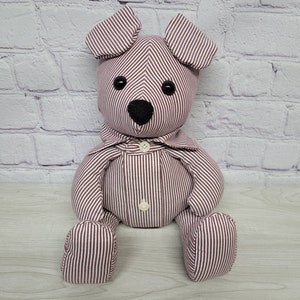 May include: A plush toy bear with a striped pattern in shades of burgundy and white. The bear has a button nose and black eyes. The bear is sitting with its legs crossed.