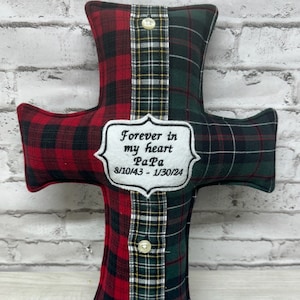May include: A red and green plaid cross-shaped pillow with a white embroidered patch that reads "Forever in my heart Papa 8/10/43 - 1/30/24".