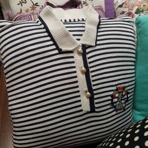 May include: A white and navy blue striped pillow with a white collar and gold buttons. The pillow has an embroidered emblem with a crown and anchor on the front.
