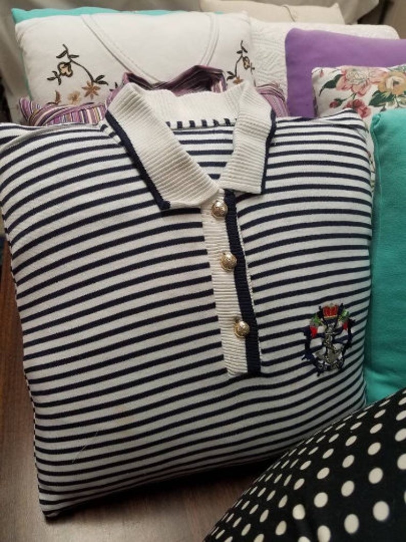 May include: A white and navy blue striped pillow with a white collar and gold buttons. The pillow has an embroidered emblem with a crown and anchor on the front.