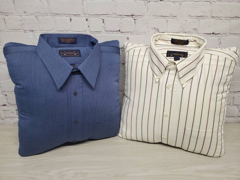 May include: Two decorative pillows shaped like shirts. One pillow is a solid blue with a white button and a pocket. The other pillow is white with thin red and white stripes and a white button.