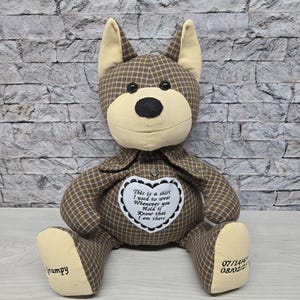 May include: A brown and tan plaid stuffed animal in the shape of a dog. The dog is wearing a shirt with a heart-shaped patch that says "This is a shirt I used to wear Whenever you hold it Know that I am there". The dog's paws are embroidered with the dates "07/14/4" and "08/02/21".