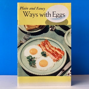 以下が含まれることがあります： 黄色い表紙に「Plain and Fancy Ways with Eggs」というタイトルが書かれたビンテージの料理本。表紙には、目玉焼きとベーコンの皿の写真が載っている。