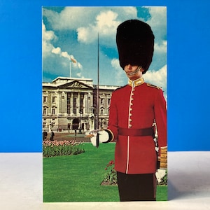 Puede incluir: Un guardia con uniforme rojo y gorro de piel de oso negro se encuentra frente al Palacio de Buckingham. El palacio es un edificio grande y blanco con muchas ventanas. Hay flores en primer plano.