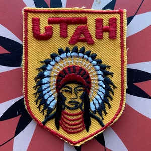 May include: A yellow and red embroidered patch with the word "UTAH" in red lettering and a Native American chief wearing a headdress.