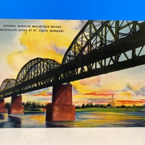 Puede incluir: Una postal vintage que representa el puente General Douglas MacArthur sobre el río Misisipi en St. Louis, Misuri. El puente es un puente de arco de acero con múltiples tramos, y la postal muestra una vista del puente al atardecer con un cielo colorido.