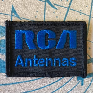 May include: A rectangular patch with the embroidered text "RCA Antennas" in blue on a black background. The patch has a black border and is likely a vintage or retro item.