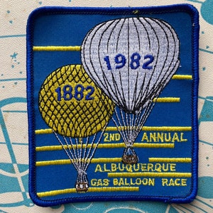 May include: Blue embroidered patch with two hot air balloons. The top balloon is white with black lines and the text "1982". The bottom balloon is yellow with black lines and the text "1882". The patch also has the text "2nd Annual Albuquerque Gas Balloon Race".