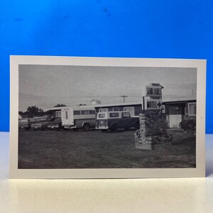 May include: Monochrome photograph of a vintage motel and vehicles. The sign reads "Sand Dunes Motel" with an arrow. Several cars and a bus are parked in front of the motel. The image is set against a blue background.