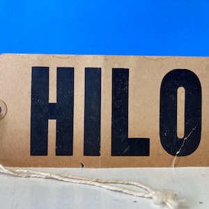 May include: A rectangular, brown paper tag with the word "HILO" printed in bold, black letters. The tag has a hole reinforced with a metal grommet and a string attached. The background is a solid blue.