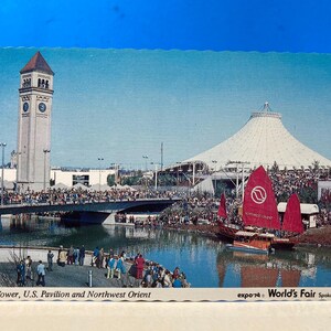 May include: Vintage postcard featuring the U.S. Pavilion and Northwest Orient at the 1974 World's Fair in Spokane, USA. The image shows a tall clock tower, a large white tent structure, and a bridge over a body of water with boats.