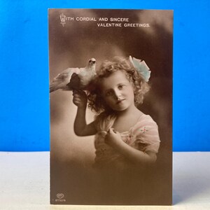 May include: Vintage sepia-toned postcard featuring a young girl holding a white dove. The girl wears a light-colored dress and has a flower in her hair. The postcard has text at the top that reads "With Cordial and Sincere Valentine Greetings."