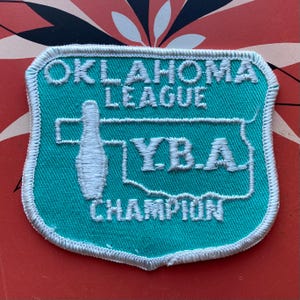 May include: A teal and white embroidered patch with the text "OKLAHOMA LEAGUE Y.B.A. CHAMPION". The patch features a bowling pin graphic and a white border.