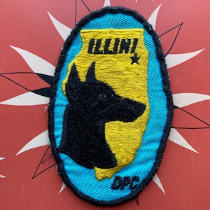 Puede incluir: Un parche bordado azul y amarillo con una silueta negra de la cabeza de un perro. El parche dice "ILLINI" y "DPC".