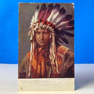 Op de afbeelding: Vintage ansichtkaart met een portret van een Indiaanse man. Hij draagt een gevederde hoofdtooi met rode en witte veren, een sjaal met patroon en een versierd kledingstuk. De ansichtkaart heeft tekst onderaan.