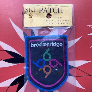 Puede incluir: Un parche de esquí de Beautiful Colorado. El parche tiene forma de escudo con fondo negro, la palabra "breckenridge" en blanco y los números "69" en verde, azul y naranja. El parche está ribeteado en azul y rosa.