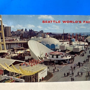 May include: Vintage postcard featuring the Seattle World's Fair. The image showcases futuristic architecture, including a large white structure and a domed building. The text "SEATTLE WORLD'S FAIR" is printed in red at the top. People are walking on the streets.