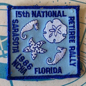 May include: Square embroidered patch with a light blue background and a dark blue border. The patch features white embroidered images of a seahorse, starfish, and sand dollar. Text includes "15th NATIONAL RETIREE RALLY SARASOTA FLORIDA 1986 NCHA".