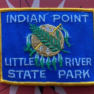 May include: A blue and white embroidered patch with yellow trim. The patch reads "Indian Point Little River State Park". The center of the patch features a stylized image of a Native American headdress.