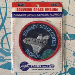 Op de afbeelding: Een ronde geborduurde patch met een zilveren spaceshuttle en een groene en blauwe aarde op een donkerblauwe achtergrond. De tekst luidt Kennedy Space Center Florida. Het is bevestigd op een witte kaart met Souvenir Space Emblem.