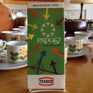 Può includere: Un opuscolo verde e bianco con il testo "Destination Expo Expo67 April 28 - October 27" e un logo per Texaco. L'opuscolo è su un tavolo con diverse tazze da tè e piattini.