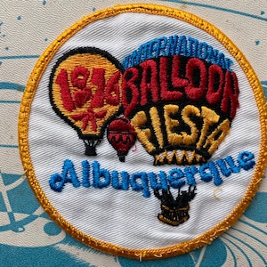 May include: A white embroidered patch with a gold border. The patch features three hot air balloons with the text "International Balloon Fiesta Albuquerque" in black and gold lettering.