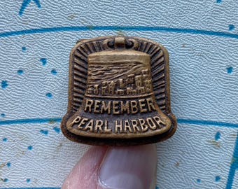 May include: A gold-colored metal pin shaped like a bell with the words "Remember Pearl Harbor" inscribed on it. The bell has a depiction of a cityscape on it.