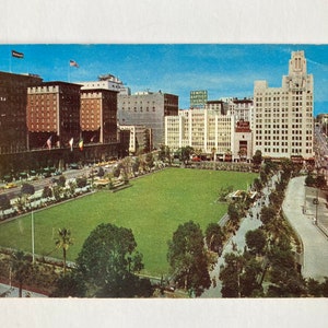 May include: A vintage postcard showing an aerial view of a city park with tall buildings in the background. The park is green and has a paved path winding through it. The buildings are mostly white and have a few red brick accents. The sky is blue and there are a few clouds.