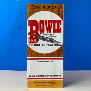 May include: A vintage city map of Bowie, Texas, with a wood-grain design. The map features the city's name in bold red letters, a knife illustration, and the text "Forged by a Century... 1882-1982." The map is a promotional item from the Bowie Chamber of Commerce.