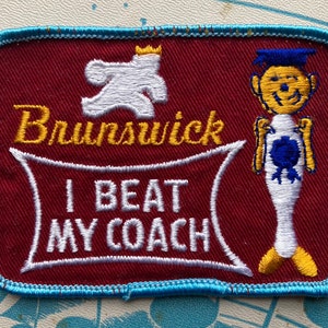 Può includere: Una toppa ricamata rossa e blu con una figura bianca che indossa una corona e il testo "Brunswick". Sotto la figura c'è uno stendardo bianco con il testo "I BEAT MY COACH". Una figura gialla che indossa un cappello di laurea blu si trova sul lato destro della toppa.