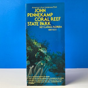 Op de afbeelding: Een brochure voor John Pennekamp Coral Reef State Park in Key Largo, Florida. De brochure toont een onderwater scène met kleurrijke vissen die zwemmen tussen koraalriffen. De tekst op de brochure luidt "America's first Undersea Park" en "John Pennekamp Coral Reef State Park".
