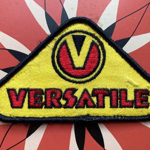 Op de afbeelding: Gele driehoekige patch met zwarte stiksels. Een gestileerde "V" in rood en zwart bevindt zich in het midden van de patch. De tekst "VERSATILE" is in rood onder de "V" geborduurd.