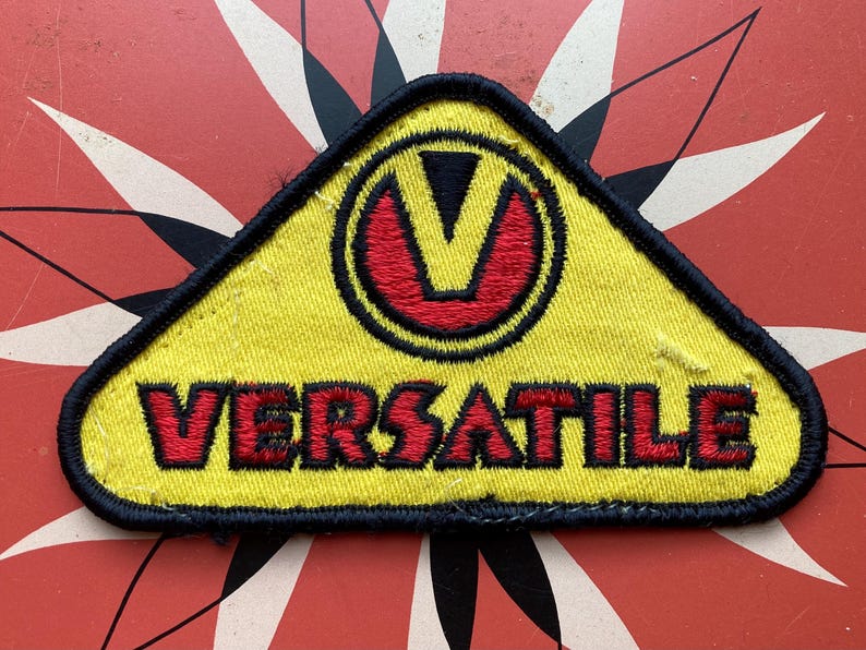 May include: Yellow triangular patch with black stitching. A red and black stylized "V" is in the center of the patch. The text "VERSATILE" is embroidered in red below the "V".