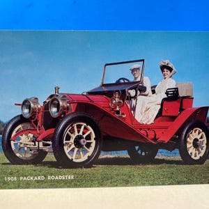 Op de afbeelding: Vintage ansichtkaart met een rode 1908 Packard Roadster met twee personen. De auto heeft witte wielen en zwarte banden. De achtergrond toont een blauwe lucht en water. De tekst "1908 Packard Roadster" staat onderaan.