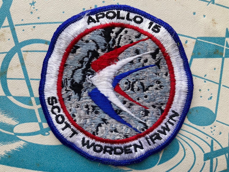 Peut inclure: Un &eacute;cusson brod&eacute; circulaire avec un fond blanc et une bordure bleue, rouge et blanche. Le centre de l'&eacute;cusson pr&eacute;sente un oiseau blanc stylis&eacute; avec des ailes rouges et bleues sur un fond gris. Le texte "APOLLO 15" est imprim&eacute; en noir en haut de l'&eacute;cusson, et le texte "SCOTT WORDEN IRWIN" est imprim&eacute; en blanc autour du bas de l'&eacute;cusson.