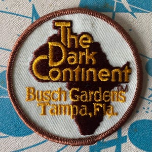 May include: A circular embroidered patch with a white background and a brown border. It features a dark brown silhouette of the African continent with yellow embroidered text that reads The Dark Continent, Busch Gardens, and Tampa, Fla.