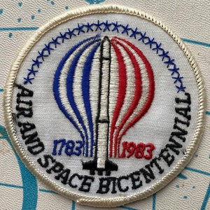 Puede incluir: Parche blanco bordado con un globo aerostático rojo, blanco y azul con un cohete en el centro. El texto alrededor del globo dice "Air and Space Bicentennial 1783-1983".