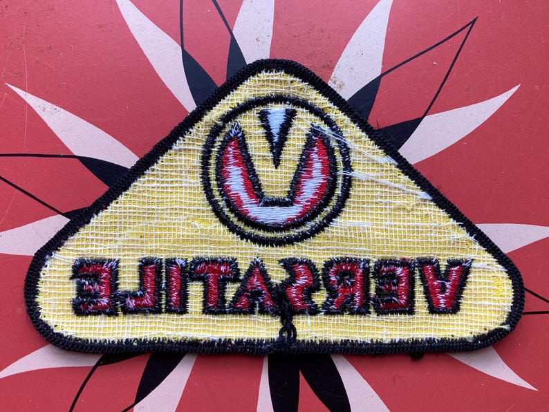 May include: Yellow triangular patch with black stitching and a red and white stylized "V" logo. The text "VIBRATE" is written in red letters below the logo.