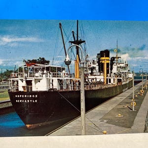 May include: Vintage postcard featuring the ship Hoperidge in Newcastle. The black and green ship is docked in a canal, with people walking along the side. The sky is blue with some clouds.