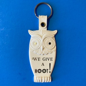 May include: White owl-shaped keychain with a metal ring. The keychain has the phrase "WE GIVE A HOOT!" in black text. The owl has detailed feather and eye designs. The background is a solid blue.